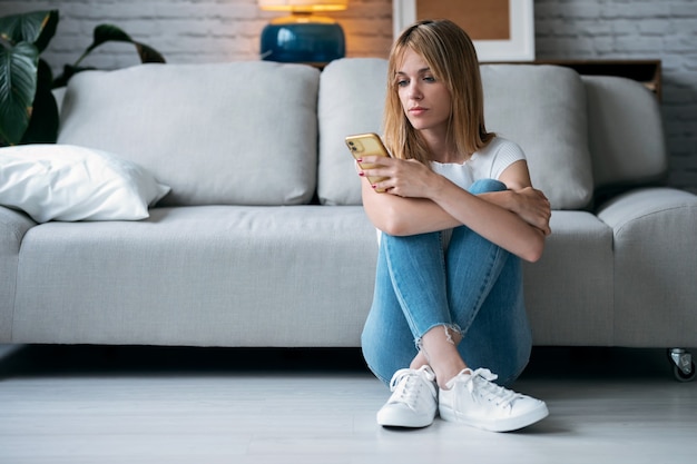 Shot of serious young woman using her mobile phone while sitting on the floor in the living room at home.