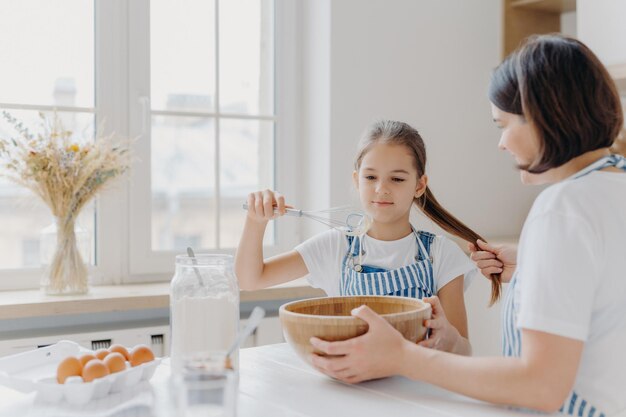 小さな子供のヘルパーのショットは、ビーターと一緒にクリームを泡立て器で焼く方法を学びます彼女のポニーテールを持っている母親からの有用なアドバイスを聞きます白いキッチンテーブルに一緒に座って家族で一緒に料理