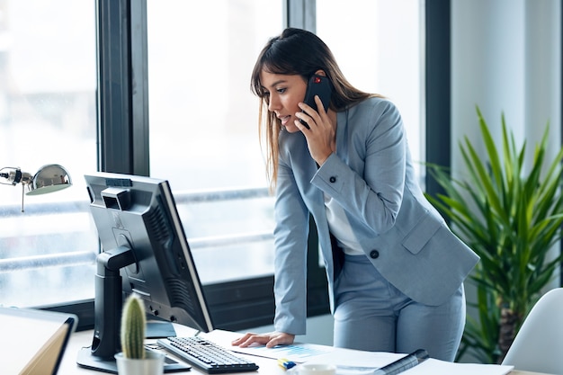 Shot of smart young businesswoman talking with mobile phone while working with computer in modern workspace.