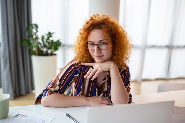 Снимка улыбающейся бизнес-женщины, работающей с ноутбуком, глядя на камеру в современном офисе стартапа