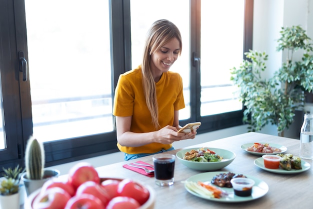 自宅で携帯電話を使用しながら健康的な食事を食べている笑顔の女性のショット。