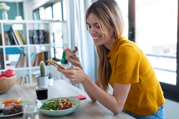 Shot of smiling woman eating healthy food while using her mobile phone at home.