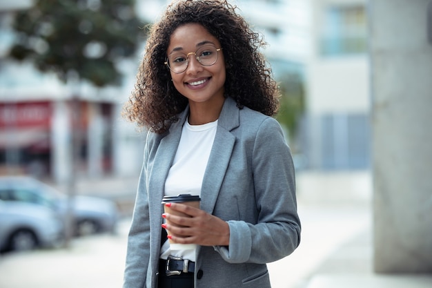 Shot of smiling young business woman drinking coffee while looking at camera in the street.