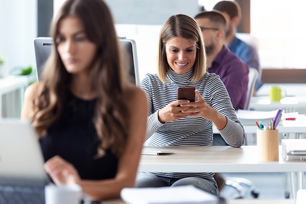 Shot of smiling young businesswoman using her smartphone while working with computer in the modern startup office.