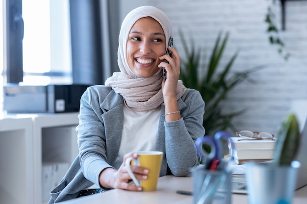 사무실에 앉아 휴대 전화로 얘기 하는 hijab을 입고 웃는 젊은 이슬람 비즈니스 여자의 쐈 어.