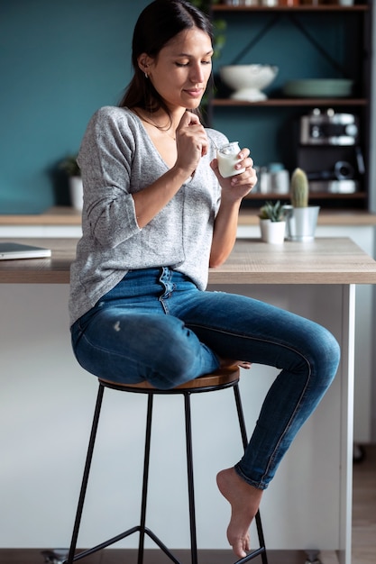 Выстрел улыбающейся молодой женщины, едящей йогурт, сидя на стуле на кухне у себя дома.