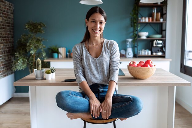 Shot of smiling young woman looking at camera while sitting on the stool in the kitchen at home.