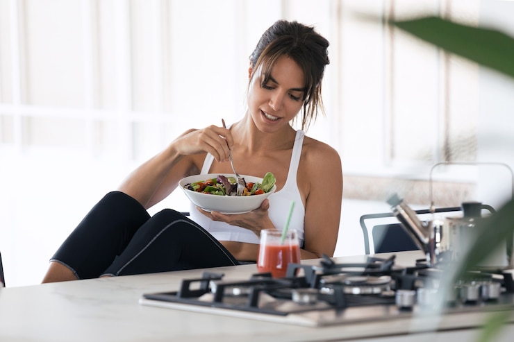 Photo shot of sporty young woman eating salad and drinking fruit juice in the kitchen at home.