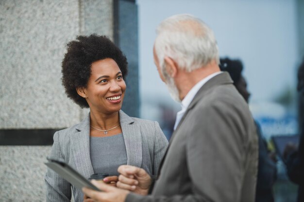 Shot of successful black businesswoman and her senior male colleague using digital tablet and having a discussion during a coffee break in front of the office building.