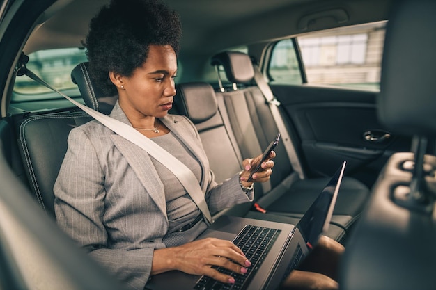 Shot of a successful black woman using smartphone while working on a laptop at backseat of a car during her business commute.