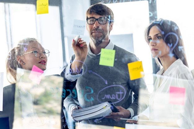 Scatto di un team aziendale di successo che scrive note alla lavagna di vetro dell'ufficio mentre discute insieme nello spazio di coworking.