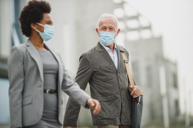 Shot of successful senior businessman and his black female colleague using protective mask and talking while walking in front of the office building during COVID-19 pandemic.