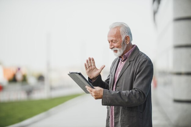 企業の建物の前でデジタルタブレットでビデオ通話を行う成功したシニアビジネスマンのショット。