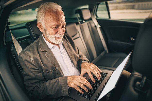 Shot of a successful senior businessman using laptop while sitting in the backseat of a car during his morning commute.