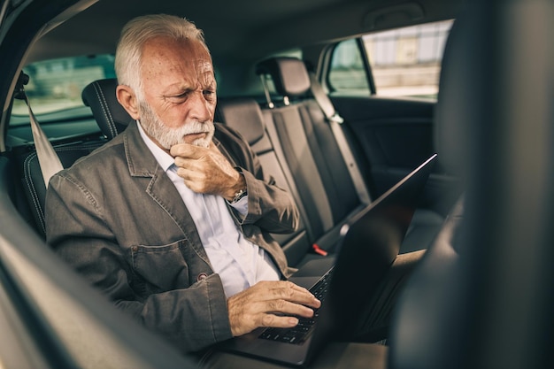 Shot of a successful senior businessman working on a laptop while sitting in the backseat of a car during his morning commute.