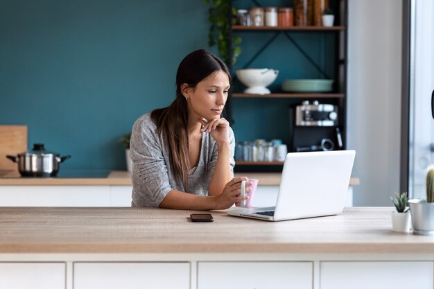 Shot of thoughtful young woman looking her laptop while holding a cup of coffee in the kitchen at home.