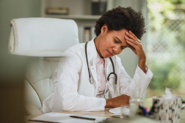 Photo shot of a tired african female doctor sitting alone at the desk in her consulting room during covid-19 pandemic.