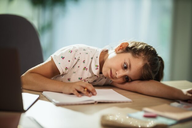 Photo shot of a tired little girl using laptop to do a online lesson at home during covid-19 pandemic.