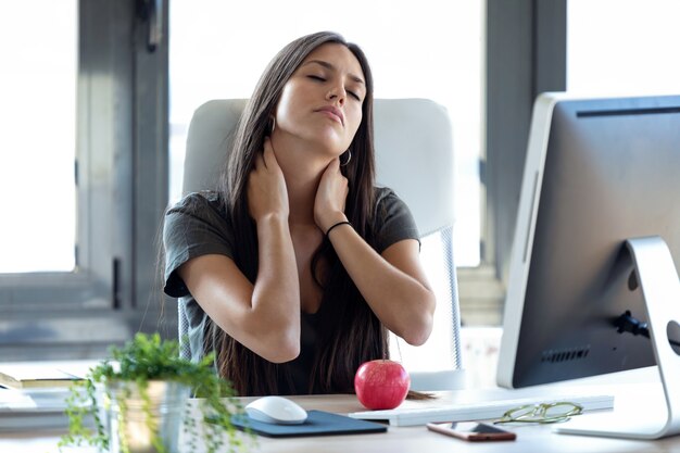 Shot of tired young business woman with neck pain while working with computer in the office.