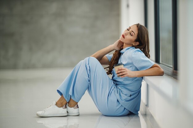 코비드-19 전염병 동안 빈 병원 복도에서 빠른 휴식을 취하는 동안 창가 근처 바닥에 앉아 있는 피곤한 젊은 간호사의 총.