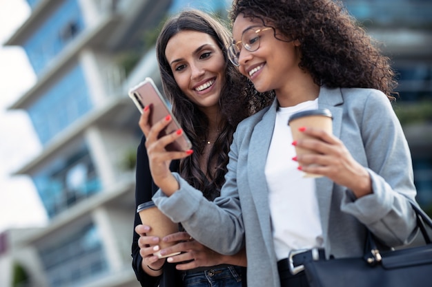 Scatto di due belle donne d'affari che usano lo smartphone mentre bevono caffè passeggiando per la città.