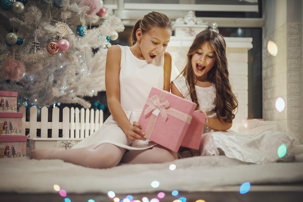 家の白いクリスマスツリーでクリスマスプレゼントを開ける2人の興奮した少女のショット。