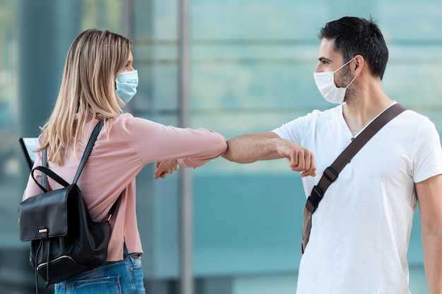 코비드-19 발병 동안 대학 캠퍼스에서 수술용 마스크를 쓰고 팔꿈치로 손을 흔드는 두 친구의 총. 사회적 거리와 새로운 정상 개념.