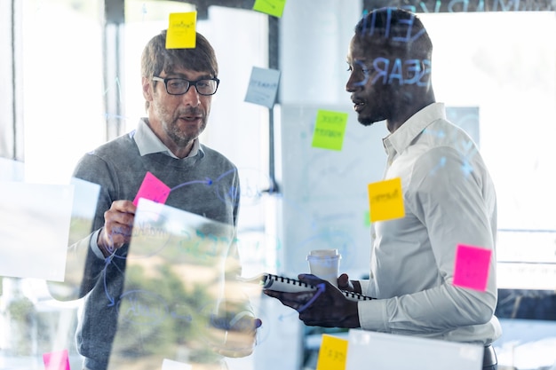 Shot of two handsome business men writing notes at office glass board while discussing together in the coworking space.