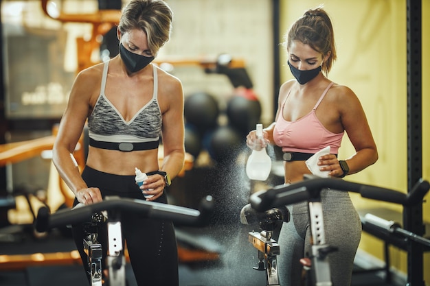 Shot of a two muscular young women with protective mask cleaning fitness gym equipment afther workout during Covid-19 pandemic.