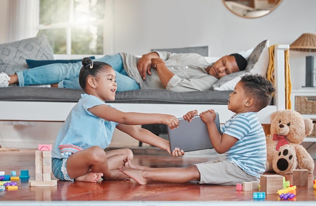 Shot of two siblings fighting over a digital tablet on the floor while their dad sleeps on the couch