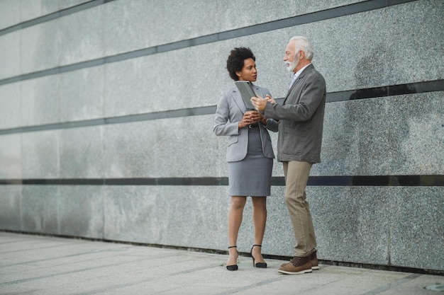 Shot of two successful multi-ethnic business people using digital tablet and having a discussion while standing against a wall of corporate building.