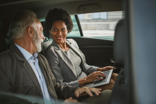 Photo shot of two successful multi-ethnic people using laptop and talk about work while sitting in the backseat of a car during their business commute.