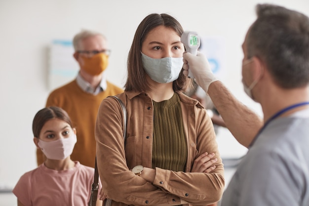 Foto colpo di medico irriconoscibile che controlla la temperatura della giovane donna che indossa la maschera e aspetta in fila in clinica o in ospedale, copia spazio