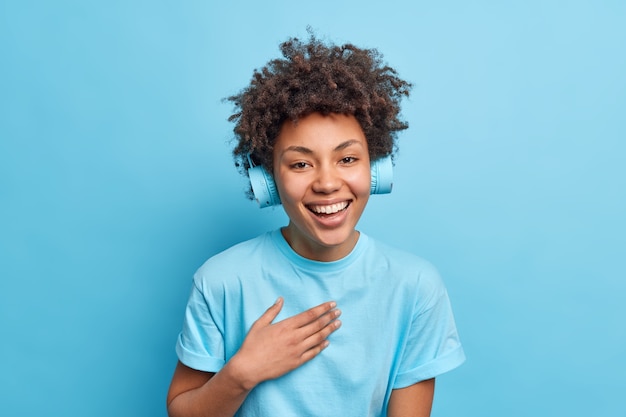 Shot van blij afro-amerikaans meisje glimlacht positief gekleed in een casual t-shirt en vermeldt muziek via een koptelefoon die geamuseerd is, poses tegen de blauwe muur. monochroom schot. positieve emoties.