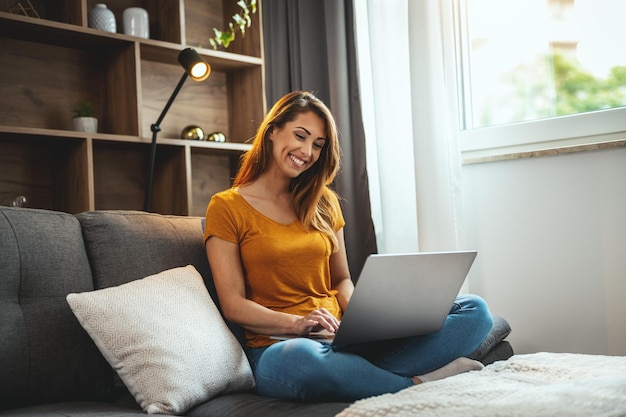 Shot van een aantrekkelijke jonge vrouw die met gekruiste benen op de bank zit en haar laptop thuis gebruikt.