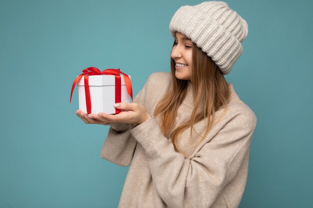 Shot van een aantrekkelijke positief lachende jonge donkerblonde vrouw geïsoleerd over een kleurrijke achtergrondmuur die een alledaagse trendy outfit draagt met een geschenkdoos en kijkt naar de zijkant. Ruimte kopiëren, mockup