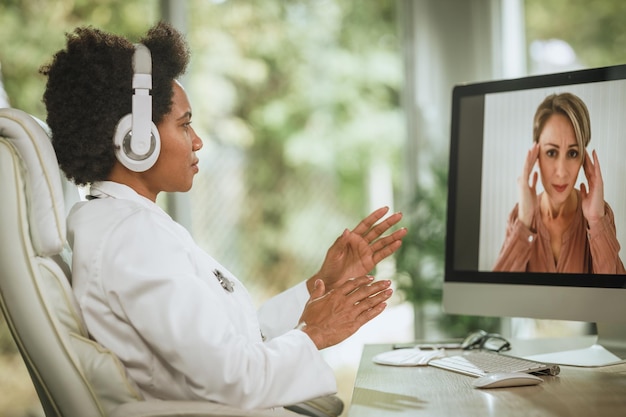 Shot van een Afrikaanse vrouwelijke arts die een videogesprek voert met de patiënt op de computer in haar spreekkamer tijdens de COVID-19-pandemie.