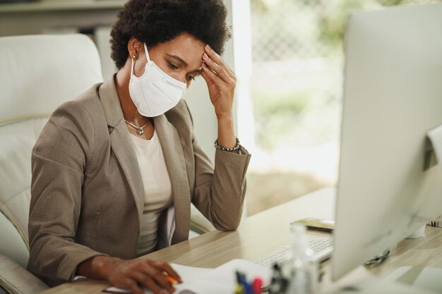 Shot van een bezorgde Afrikaanse zakenvrouw met beschermend N95-masker die alleen in haar kantoor zit en op de computer werkt tijdens de COVID-19-pandemie.