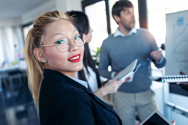 Foto shot van een elegante jonge zakenvrouw die naar de camera kijkt terwijl haar collega's het project in een coworking-plek uitleggen.