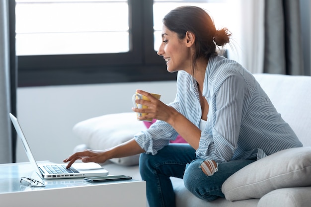 Shot van een gelukkige zakenvrouw die op afstand thuis werkt met een laptop en koffie drinkt terwijl ze op de bank zit.
