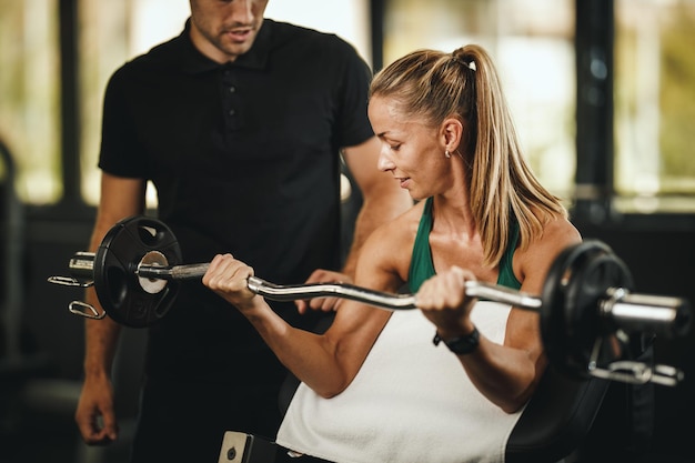 Shot van een gespierde jonge vrouw in sportkleding die traint met een personal trainer in de sportschool. Ze pompt haar bicepsspier met zwaar gewicht op.