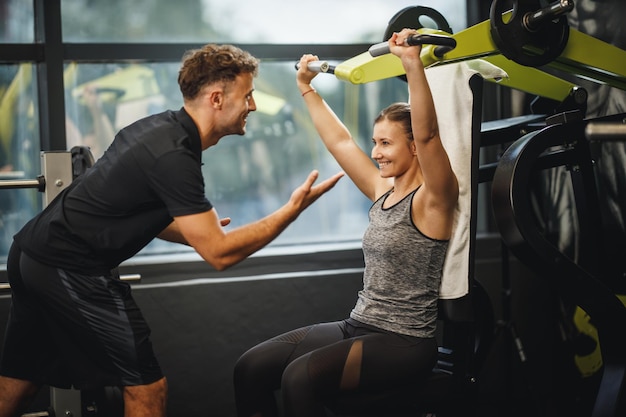 Shot van een gespierde jonge vrouw in sportkleding die traint met een persoonlijke trainer op de sportschoolmachine. Ze pompt haar schouderspieren op met zwaar gewicht.