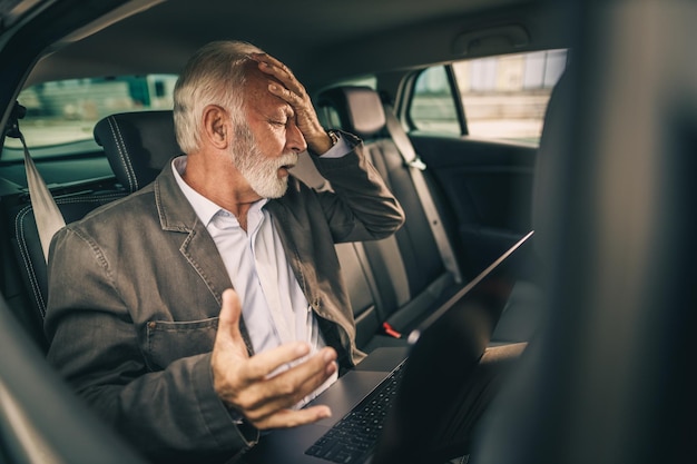 Shot van een gestresste senior man die laptop gebruikt terwijl hij op de achterbank van een auto zit tijdens zijn zakelijke woon-werkverkeer.