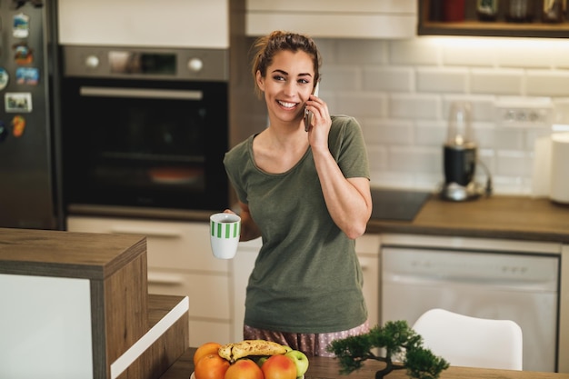 Shot van een jonge vrouw die koffie drinkt terwijl ze haar smartphone thuis gebruikt.