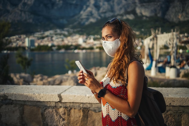 Shot van een jonge vrouw met beschermend masker die smartphone gebruikt en geniet van een vakantie op het strand tijdens de COVID-19.