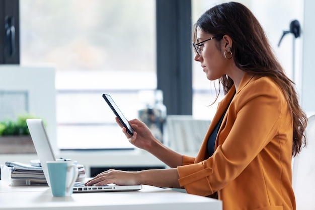 Shot van een jonge zakenvrouw die haar mobiele telefoon gebruikt terwijl ze met een laptop op kantoor werkt.