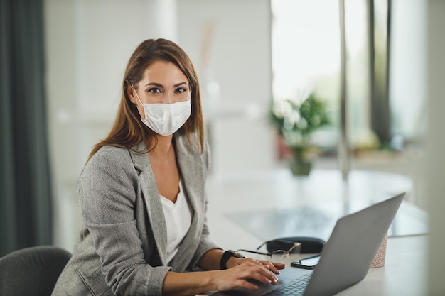 Shot van een jonge zakenvrouw met beschermend masker die alleen in haar thuiskantoor zit en op een laptop werkt tijdens de COVID-19-pandemie. Camera kijken.