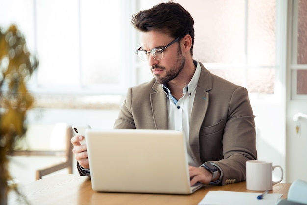 Shot van een knappe jonge zakenman die zijn smartphone gebruikt terwijl hij met een laptop op kantoor werkt.