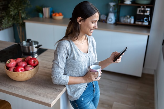 Shot van een lachende jonge vrouw die haar mobiele telefoon gebruikt terwijl ze thuis een kopje koffie drinkt in de keuken.