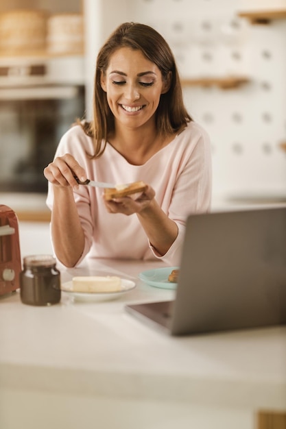 Foto shot van een lachende jonge vrouw die het ontbijt voor zichzelf klaarmaakt, boter op toast smeert in haar keuken en een laptop gebruikt.